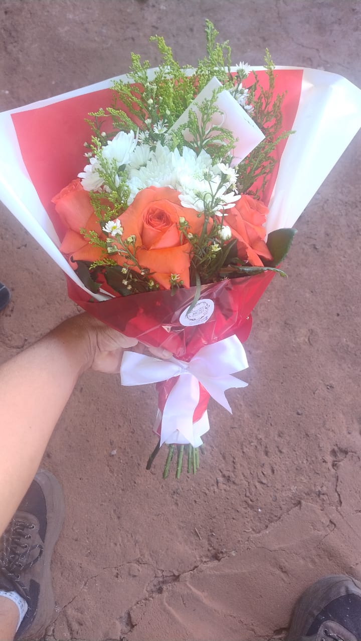 mão segurando um buquê de rosas laranjas com margaridas brancas e folhagens, embalado em papel vermelho e bran