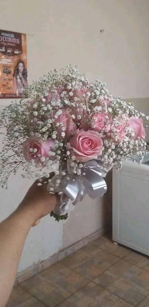 mão segurando um buquê de rosas cor-de-rosa com gipsofila e laço de fita prateada num ambiente interno