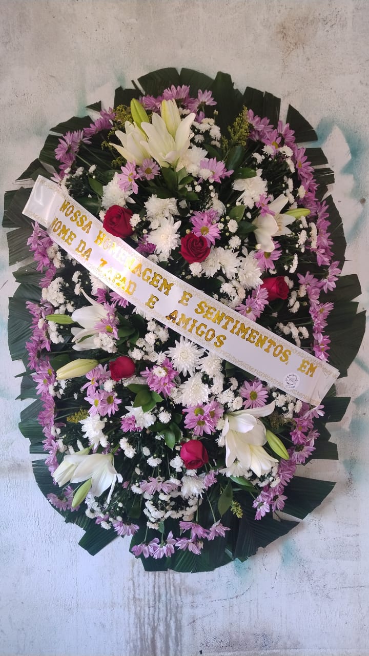coroa de flores funerária oval com lírios brancos, rosas vermelhas, margaridas roxas e folhas verdes sobre parede clara,
