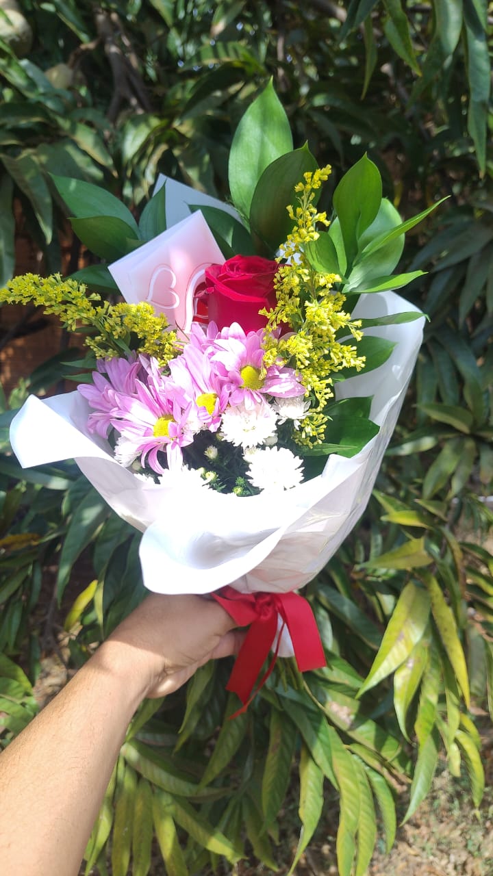mão segurando um buquê de flores com rosa vermelha, margaridas rosadas e brancas embrulhado em papel branco co