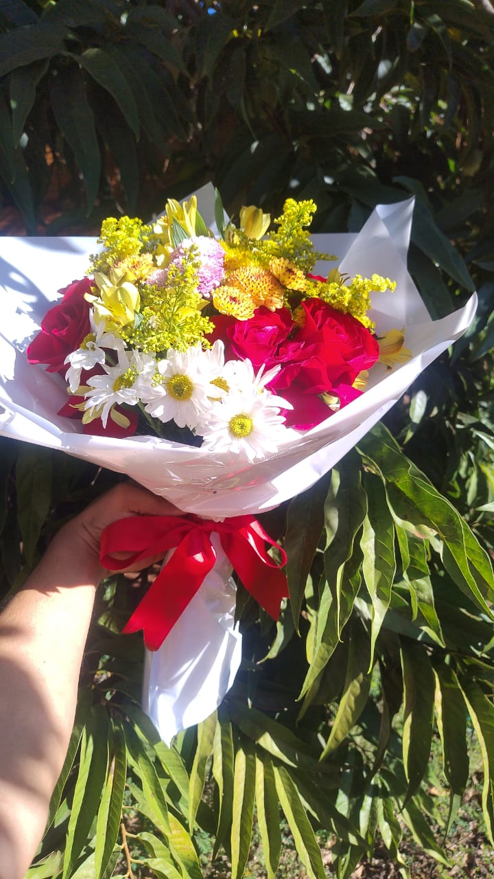 mão segurando um buquê de flores coloridas com rosas vermelhas, margaridas e fita vermelha embrulhado em papel