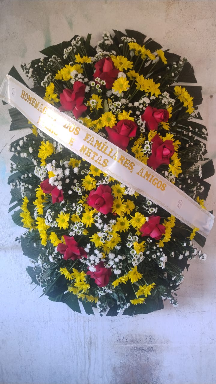 coroa de flores com rosas vermelhas, margaridas amarelas e brancas com faixa de homenagem pendurada na parede clara