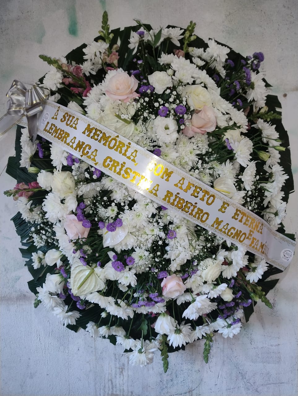 coroa de flores fúnebre circular com flores brancas, rosas cor-de-rosa e flores roxas, com faixa dourada de homenagem