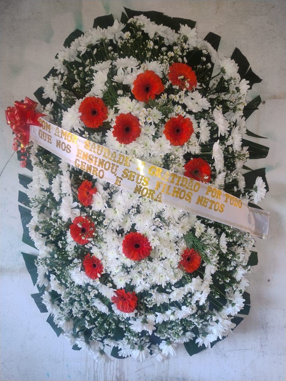 coroa de flores funerária com margaridas brancas, gerberas vermelhas e fita com mensagem de homenagem
